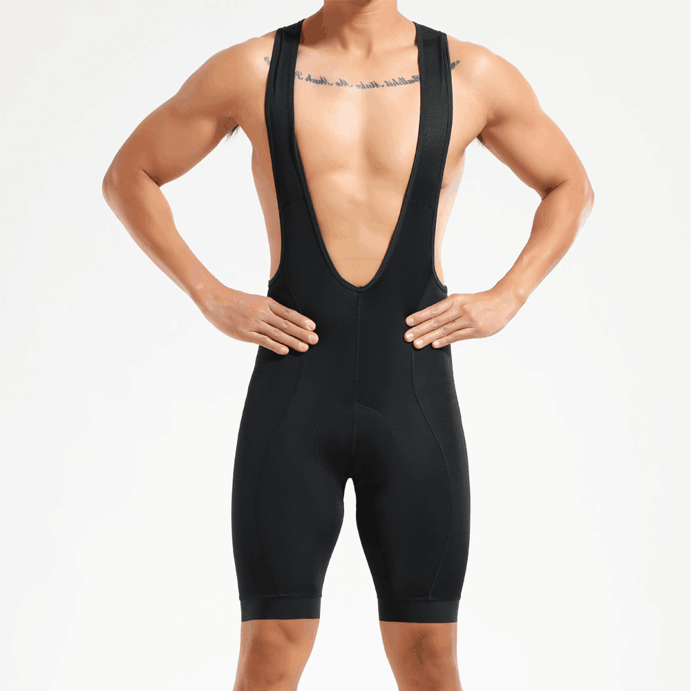 A man models Pro Team Cycling Bib Shorts, featuring suspenders and a dual density chamois pad for performance comfort. He poses against a plain white background with hands on hips, showcasing his tattoos across the upper chest.