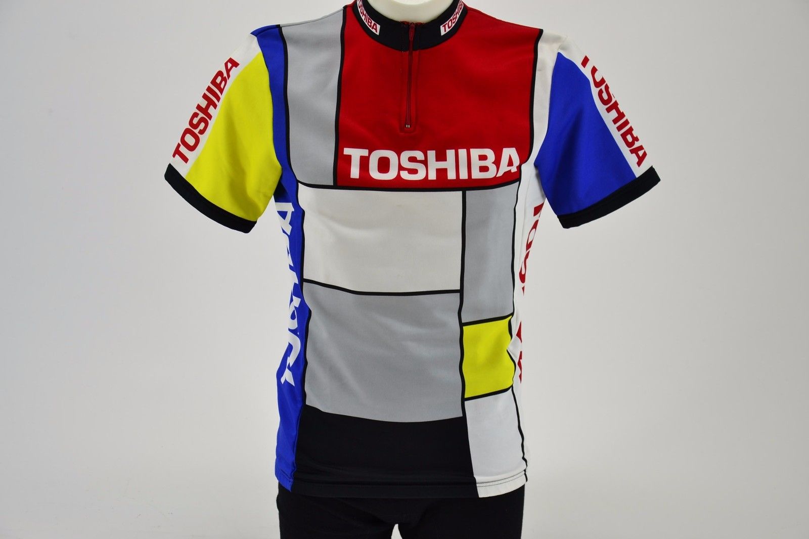 The Toshiba Look 1989 Retro Cycling Jersey features a Mondrian-inspired design with vibrant red, blue, yellow, black, and white blocks. TOSHIBA is prominently displayed in classic 1989 Tour de France styleβwhite on a red chest and striking red on white sleeves.