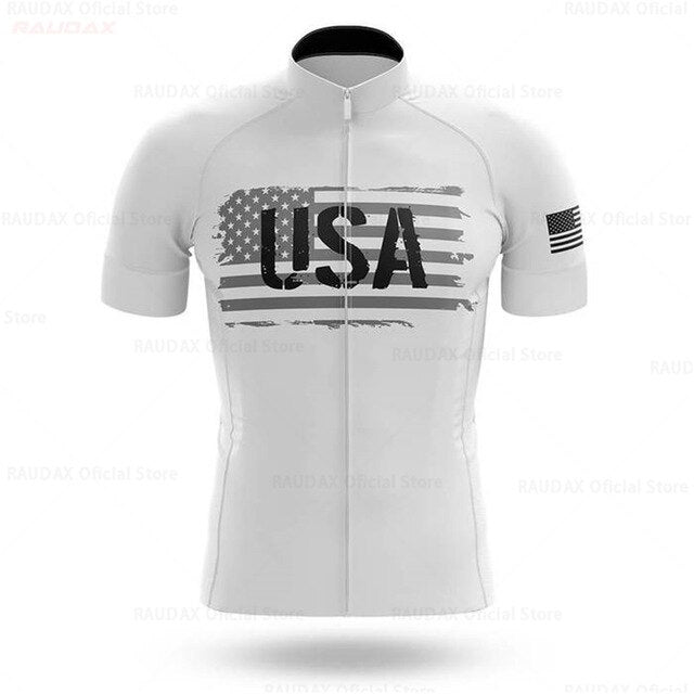 The USA White Cycling Team Jersey is a short-sleeve performance jersey made from lightweight polyester, showcasing a large distressed American flag and USA text on the front, with another flag on the left sleeve. It features a convenient front zipper.