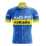 The Maes-Pils Rokado Retro Cycling Jersey, crafted from lightweight polyester, features a blue design with yellow accents. MAES Pils is displayed in white and rokado in black on the chest, while yellow trim highlights the sleeves and collar.