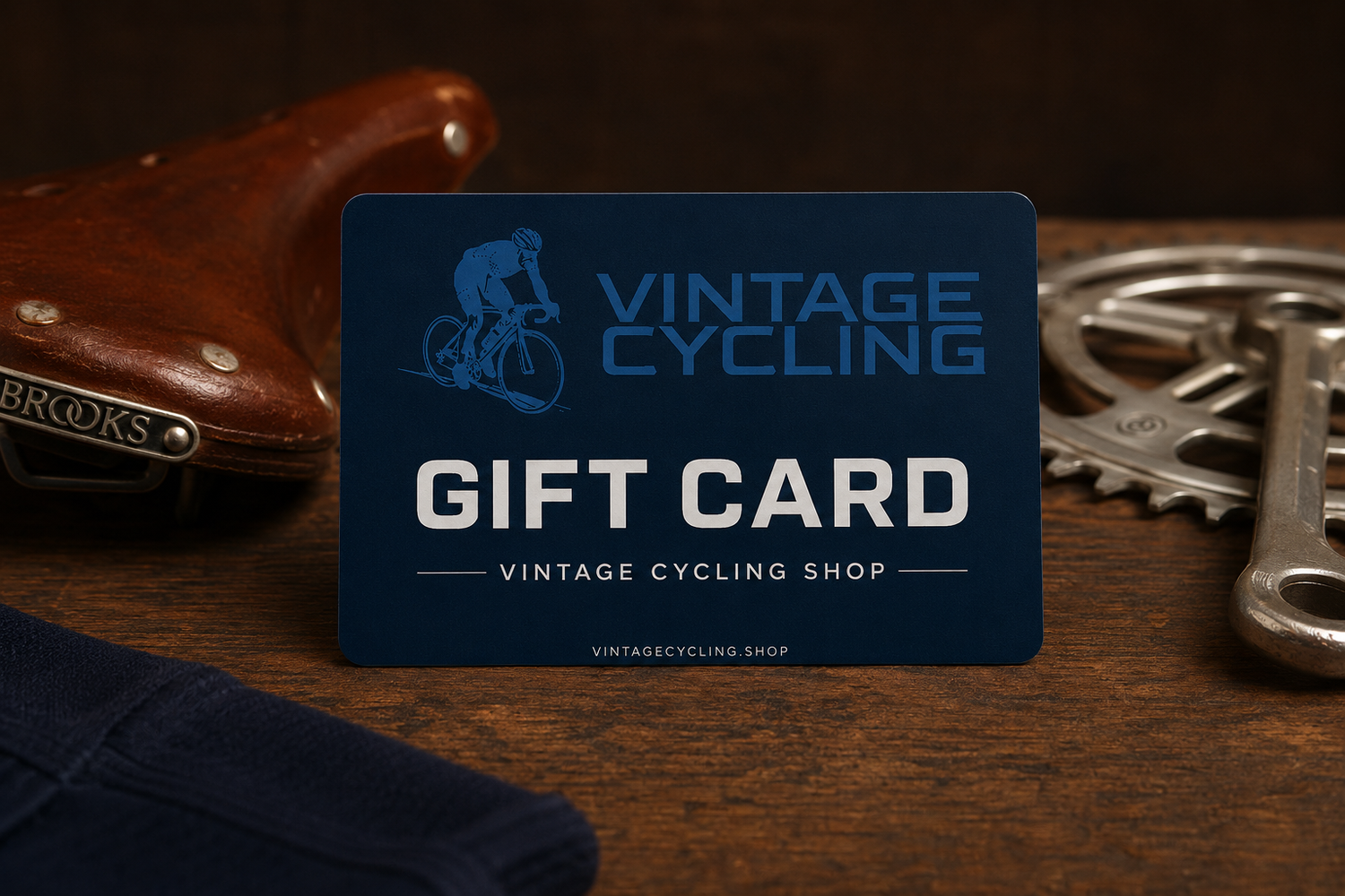 A Vintage Cycling Shop Gift Card in blue is displayed on a wooden surface, accompanied by a brown leather bicycle saddle, a silver bike gear, and a navy cloth.