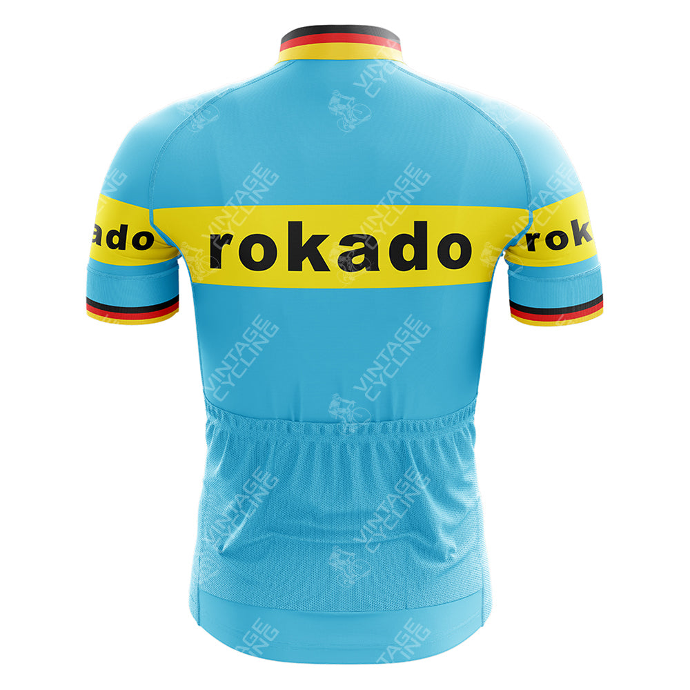 The image features the Boule dOr Sunair Retro Short Sleeve Cycling Set, a vintage-style jersey in light blue with yellow horizontal chest and arm stripes. Rokado is printed in black, with red, black, and yellow bands on the collar and arm edges.