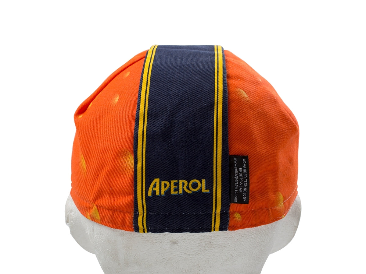 The Aperol Retro Cycling Cap features an orange vintage style with a black and yellow central stripe and bold APEROL in yellow letters. It sits on a white mannequin head, displaying a classic retro design with yellow spots.