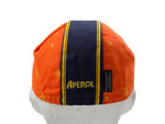 The Aperol Retro Cycling Cap features an orange vintage style with a black and yellow central stripe and bold APEROL in yellow letters. It sits on a white mannequin head, displaying a classic retro design with yellow spots.