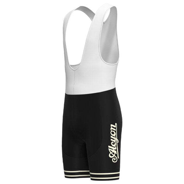 The Paris-Roubaix Retro Cycling Jersey Set features black and white cycling bib shorts with white straps for performance. Alcyon appears in bold cursive on the black lower section with two white stripes, and a breathable gel seat pad enhances comfort. The upper section forms comfortable shoulder straps.