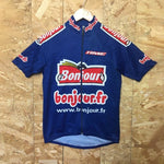 The Bonjour Retro Cycling Jersey, made of lightweight polyester, showcases a blue design with a red and white bonjour.fr logo on the front and sleeves. It hangs on a wooden hanger against a textured wall, highlighting its performance style.