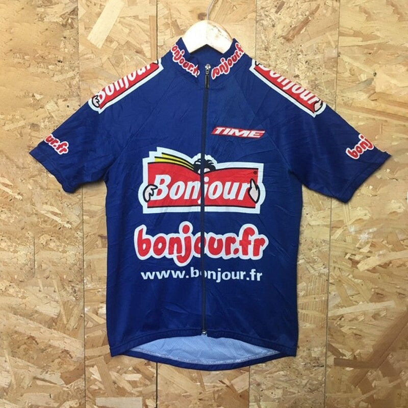 The Bonjour Retro Cycling Jersey, made of lightweight polyester, showcases a blue design with a red and white bonjour.fr logo on the front and sleeves. It hangs on a wooden hanger against a textured wall, highlighting its performance style.